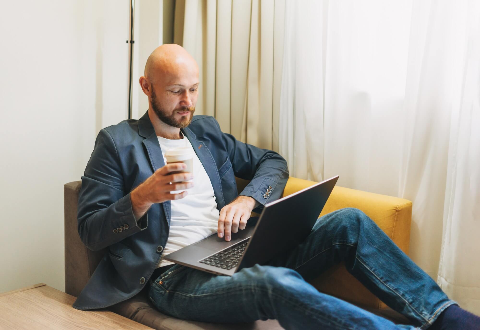 Adult bald bearded man in blue jacket with coffee working on laptop in hotel room